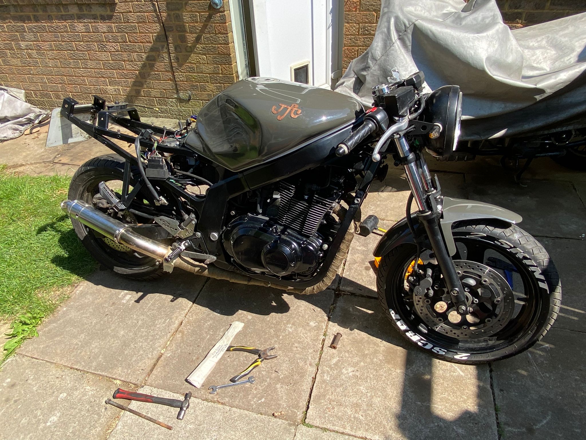 Suzuki GS500F mid-build on the driveway with JFC tank logo, no seat, tools laid out – work in progress